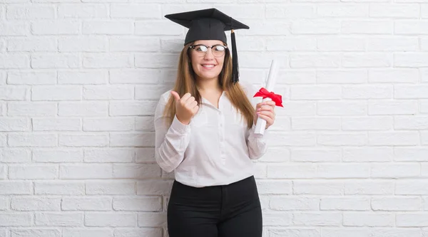 穿白砖墙的年轻成年妇女戴着研究生帽持有学位快乐的大微笑做 拇指与手指 优秀的标志 — 图库照片