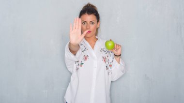 Genç Yetişkin kadın açık eliyle dur işareti ile ciddi ve kendine güvenen ifade, savunma hareketi yaparken taze yeşil elma yeme gri grunge duvarın üzerinden
