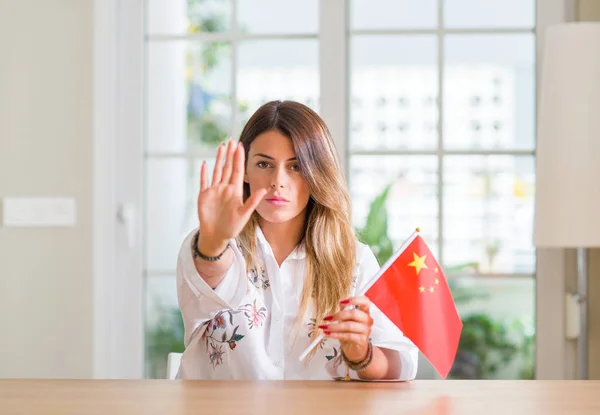 Young woman at home holding flag of China with open hand doing stop sign with serious and confident expression, defense gesture