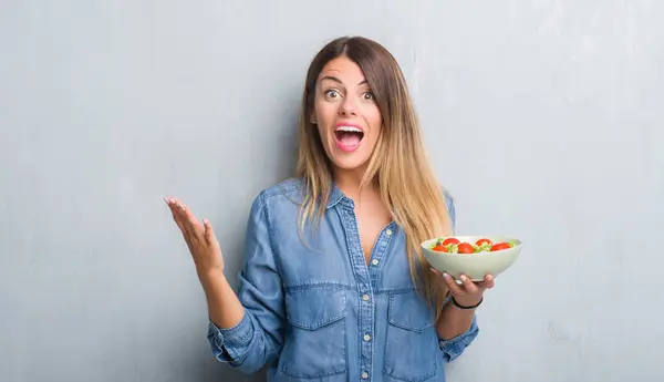 灰色垃圾墙上的年轻成年妇女吃健康的西红柿沙拉非常高兴和兴奋 优胜者表达庆祝胜利尖叫与大微笑和举手 — 图库照片