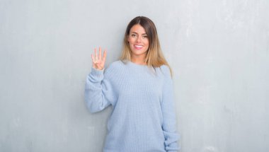 Gri grunge duvar gösterilen ve parmakları ile işaret kış kıyafeti giymiş genç yetişkin kadın kendinden emin ve mutlu gülümseyerek süre dört numara.