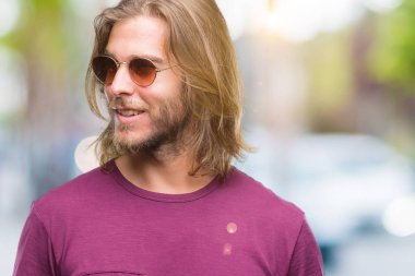 Genç yakışıklı adam uzun saçlı uzak tarafına yüzünde, doğal ifade gülümseme ile seyir izole arka plan üzerinde güneş gözlüğü takıyor. Kendine güvenen gülüyor.