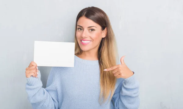 Young adult woman over grey grunge wall holding blank card with surprise face pointing finger to himself