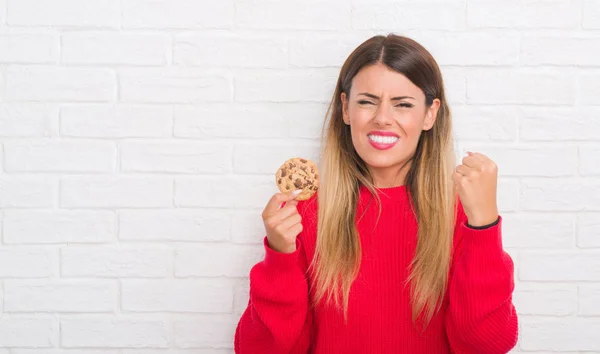 白いレンガに若い成人女性壁腹のチョコレート チップ クッキーを食べるとイライラ叫び狂気と上げられた手で 叫んで 怒りと怒りの概念 — ストック写真