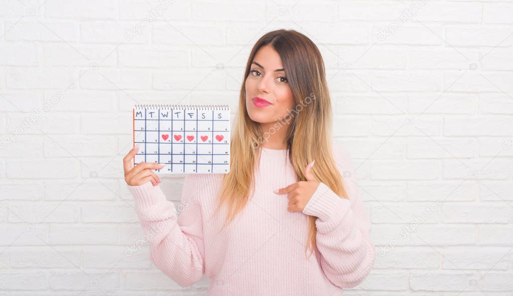Young adult woman over white brick wall holding period calendar with surprise face pointing finger to himself