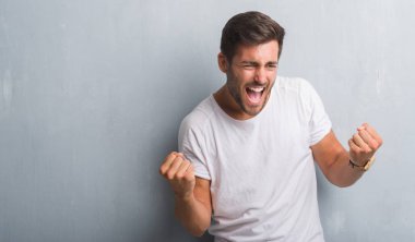 Gülümseyen ve başarı için çığlık yakışıklı genç adam gri grunge duvar çok mutlu ve heyecanlı kaldırdı, silah ile kazanan jest yapıyor. Kutlama kavramı.