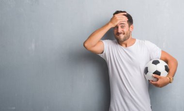 Yakışıklı genç adam kafasına, el ile vurguladı futbol futbol topu tutan gri grunge duvarın üzerinden şok utanç ve sürpriz suratla, öfkeli ve sinirli. Korku ve hata için üzgün.