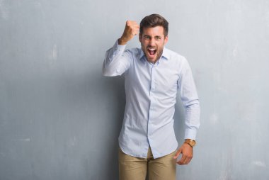 Yakışıklı genç iş adamı gri grunge üzerinde giyen şık gömlek öfkeli ve sinirli sinirli ve öfke ile bağırarak süre öfkeli yumruk yükselterek duvar. Öfke ve agresif kavramı.