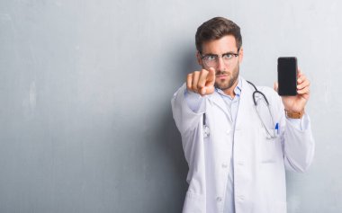 Yakışıklı genç adam akıllı telefon kamera ve size, el işareti, pozitif ve kendinden emin bir jest ön parmak ile işaret tutan gri grunge duvarın üzerinden doktor