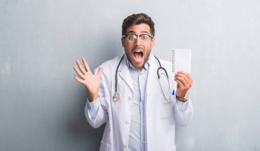 Yakışıklı genç doktor adam gri grunge duvar büyük gülümseme ile çığlık zaferini kutluyor ve ellerini kaldırdı boş defter çok mutlu ve heyecanlı, kazanan ifade holding