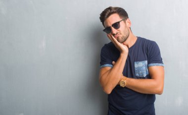 Yakışıklı genç gri grunge üzerinde duvar düşünme seyir yorgun ve çapraz kolları olan depresyon sorunları ile sıkılmış giyen güneş gözlüğü.