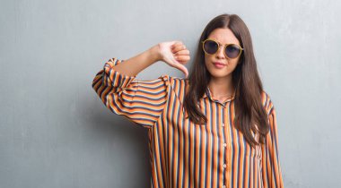 Grunge gri duvar retro güneş gözlüğü ve kızgın yüzü, eksi işareti aşağı antipati başparmak ile gösterilen, ret kavramı ile kıyafet giyen genç esmer kadın