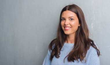 Grunge gri duvar ayakta ve diş gösterilen kendine güvenen bir gülümsemeyle gülümseyerek mutlu bir yüz ile genç esmer kadın