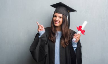 Grunge gri duvar giymiş mezunu genç esmer kadın holding derecesi bir fikirle sürpriz üniforma veya gülen yüz, bir numara ile işaret parmağı soru