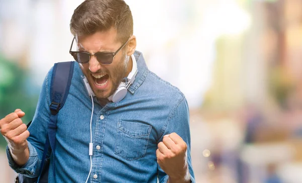 Üzerinde kulaklık ve sırt çantası giyen genç yakışıklı turist adam arka plan çok mutlu ve heyecanlı kaldırdı, silah ile kazanan jest yapıyor gülümseyen ve başarı için çığlık izole. Kutlama kavramı.