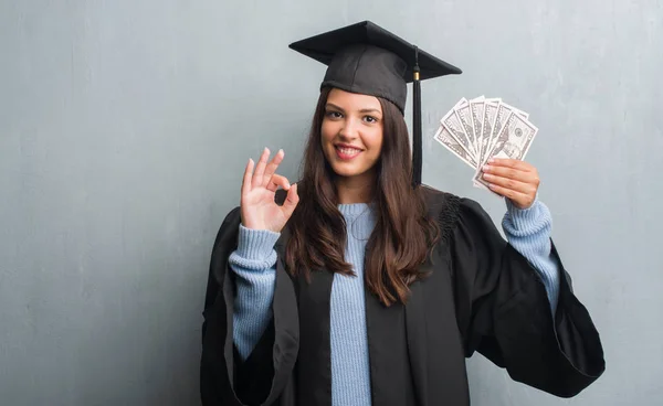 グランジの若いブルネットの女性灰色の壁 優秀なシンボルの指で サインをしてドルを保持している大学院の制服を着て — ストック写真
