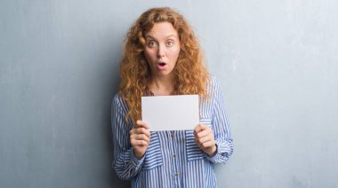 Gri grunge duvar holding boş kartı bir sürpriz yüz, korkuyor ve heyecanlı korku ifadesi ile şok korkmuş genç Kızıl saçlı kadın