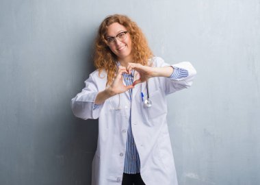 Genç redhead doktor kadın kalp simgesi ve elleriyle şekil gösterilen aşık gülümseyen bir palto giyiyor gri grunge duvarın üzerinden. Romantik kavramı.