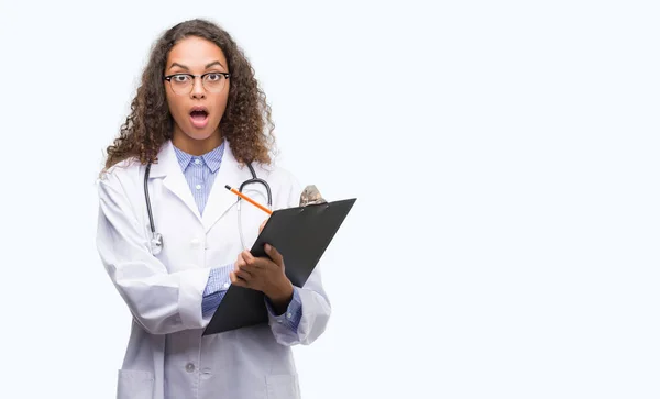 年轻的西班牙裔医生的女人拿着剪贴板吓坏了惊讶的脸 害怕和兴奋的表情恐惧表达 — 图库照片
