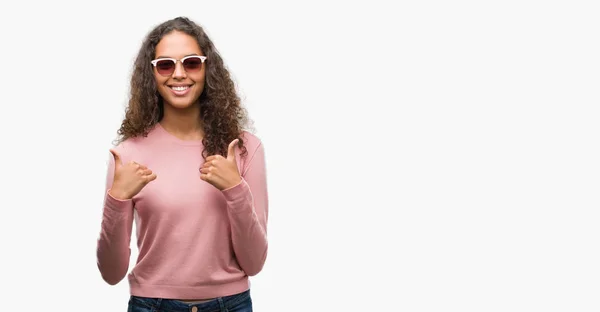 美丽的年轻西班牙裔妇女戴着太阳镜成功的标志做积极的手势与手 大拇指向上微笑和快乐 看着带着欢快表情的相机 胜利者的手势 — 图库照片
