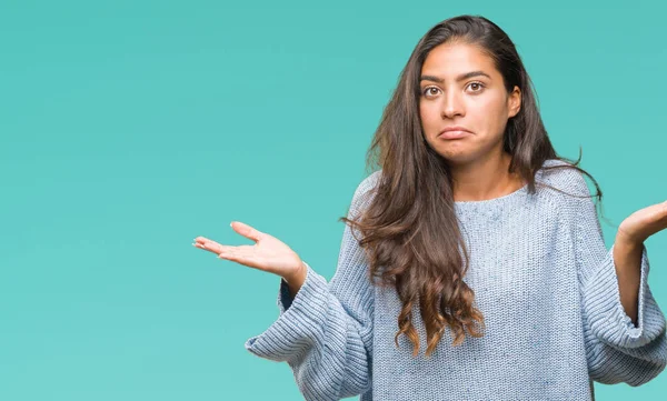 İzole arka plan clueless ve şaşkın bir ifadeyle kollar ve eller kaldırdı üzerinde kış kazak giyen genç güzel Arap kadın. Şüphe kavramı.