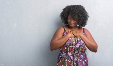 Gri grunge duvar renkli giyen genç Afro-Amerikan kadın elbise göğüs kapalı gözlerle ellerinde ve yüzünde minnettar jest ile gülümseyen. Sağlık kavramı.