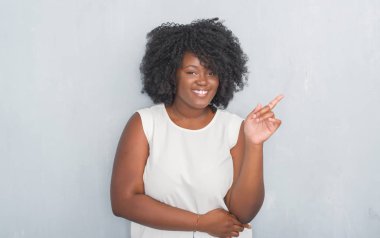 Kameraya bakarak yan el ve parmak ile işaret eden genç Afro-Amerikan kadın yüzünde kocaman bir gülümseme ile gri grunge duvarın üzerinden.