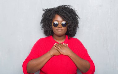 Genç Afro-Amerikan artı boyutu kadın kapalı gözler ile göğüs ellerinde ve yüzünde minnettar jest ile gülümseyen retro güneş gözlüğü takıyor gri grunge duvarın üzerinden. Sağlık kavramı.