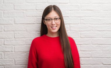 Genç Çinli kadın ayakta ve diş gösterilen kendine güvenen bir gülümsemeyle gülümseyerek mutlu bir yüz ile tuğla duvarın üzerinden