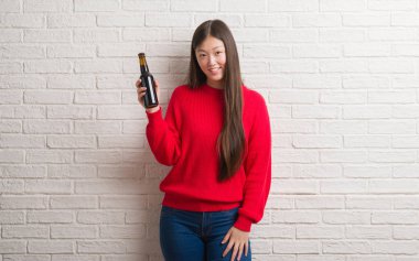 Genç Çinli kadın ayakta ve diş gösterilen kendine güvenen bir gülümsemeyle gülümseyerek mutlu bir yüz ile bira içme tuğla duvarın üzerinden