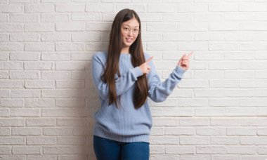 Genç Chinise kadın gülümseyerek ve iki elleri ve parmakları ile tarafına işaret kamera bakıyor beyaz tuğla duvarın üzerinden.