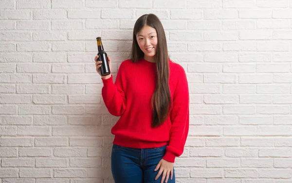 Genç Çinli kadın ayakta ve diş gösterilen kendine güvenen bir gülümsemeyle gülümseyerek mutlu bir yüz ile bira içme tuğla duvarın üzerinden