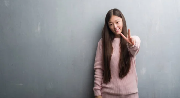 年轻的中国女人在灰色的墙上微笑着看着相机 露出手指做着胜利的手势 第二个 — 图库照片