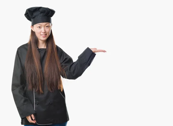 年轻的中国妇女在孤立的背景下穿着厨师制服微笑着欢快地呈现和指着手掌看着相机 — 图库照片