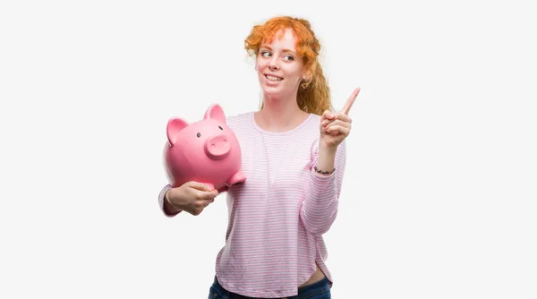 Junge Rothaarige Frau Hält Sparschwein Sehr Glücklich Und Zeigt Mit — Stockfoto