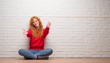 Sarılmak için kollarını açarak gülümseyerek kameraya bakarak tuğla duvar üzerine oturan genç Kızıl saçlı kadın. Neşeli ifade kucaklayan mutluluk.