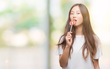Genç Asyalı kadın acımak yüz ciddi düşünüyorum üstünde kendine güvenen bir ifade ile izole arka plan üzerinde lolipop şeker yemek