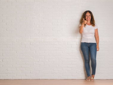 Orta yaş İspanyol kadın beyaz tuğla üzerinde duran kocaman bir gülümseme ile ok işareti yapıyor mutlu duvar, parmaklarıyla, mükemmel işaret parmak