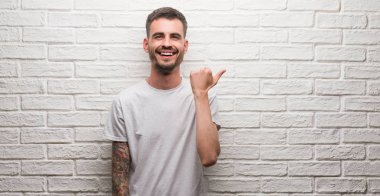 Genç yetişkin adam işaret ve başparmak kadar mutlu yüz gülümseyen tarafı ile gösterilen beyaz tuğla duvarın üzerinden ayakta