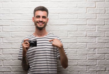 Genç adam kendisi için sürpriz yüz işaret parmağı ile beyaz tuğla duvarın üzerinden vintage fotoğraf makinesi ayakta tutan