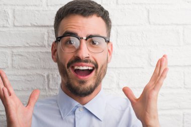 Beyaz tuğla üzerinde duran genç yetişkin iş adamı çok mutlu ve heyecanlı, kazanan ifade büyük gülümseme ile çığlık zaferini kutluyor duvar ve ellerini kaldırdı