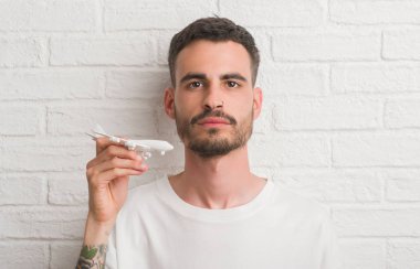 Genç yetişkin adam akıllı yüz ciddi düşünüyorum üstünde kendine güvenen bir ifade ile beyaz uçak holding tuğla duvarın üzerinden