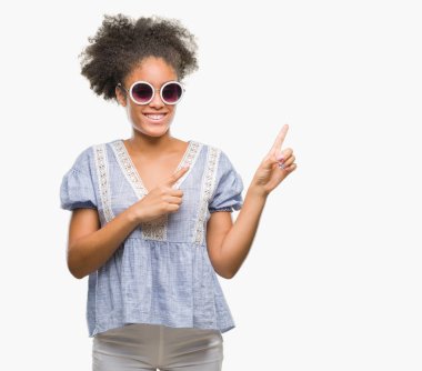 Genç afro Amerikalı kadın gülümseyerek ve iki elleri ve parmakları ile tarafına işaret kamera bakarak izole arka plan üzerinde gözlük.