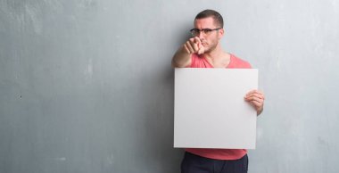 Beyaz delikanlı kamera ve size parmak ile işaret boş bir afiş tutarak gri grunge duvarın üzerinden, el işareti, pozitif ve kendinden emin bir jest ön