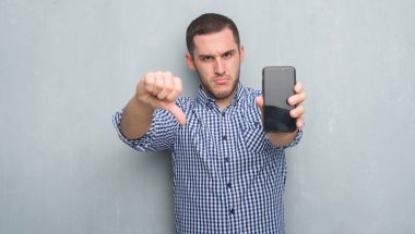 Gri grunge üzerinde beyaz delikanlı duvar boş ekran smartphone kızgın yüzü, eksi işareti aşağı antipati başparmak ile gösterilen, ret kavramı ile gösterilen