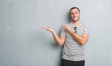 Hayret ve el ile sunulması ve parmak ile işaret ederken kameraya gülümseyen gri grunge duvarın üzerinden beyaz delikanlı.