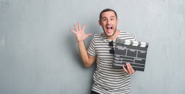 Genç beyaz adam gri grunge duvar holding film üzerinde çok mutlu ve heyecanlı, kazanan ifade büyük gülümseme ile çığlık zaferini kutluyor fıçı tahtası ve ellerini kaldırdı