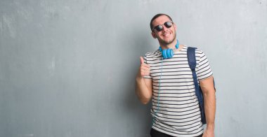 Ok işareti, başparmak ile parmak, mükemmel işareti yapıyor kocaman bir gülümseme ile genç beyaz öğrenci adam gri grunge duvar giyen kulaklık ve sırt çantası mutlu