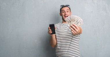 Dolar ve smarphone çok mutlu ve heyecanlı, kazanan ifade ile çığlık zaferini kutluyor büyük holding gri grunge duvarın üzerinden beyaz delikanlı gülümse ve el kaldırdı