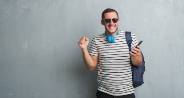 Duygu tezahürat Genç beyaz öğrenci adam gurur çığlık ve zafer ve başarı çok heyecanlı, kutluyor smartphone kullanarak kulaklık giyen gri grunge duvarın üzerinden